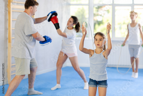 Girl jumps on skipping rope during family training session while mom and dad practice boxing and kickboxing techniques