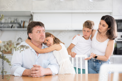 Offended father sitting at the kitchen table being hugged by little daughter, his wife and son looking at them with empathy photo