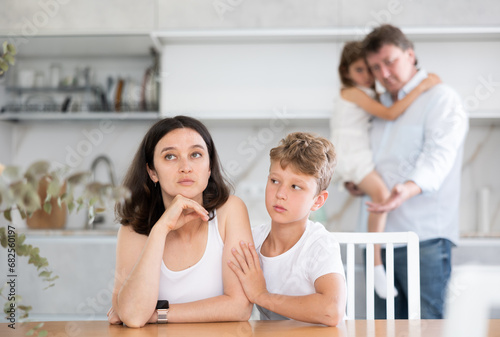 Frustrated wife sitting at kitchen table during family quarrel, with angry husband standing behind her. Children are worried about parents quarrel. Son hugs dad, daughter gently leaned against sad mom