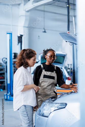 Licensed mechanic helping client with car maintenance in auto repair shop. Employee in garage facility looking over automobile parts with woman, mending her vehicle brakes during inspection