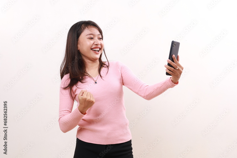Young asian woman celebrating her victory with fist hand while holding a cell phone. Isolated on white background Young asian woman celebrating her victory with fist hand while holding a cell phone. Isolated on white background