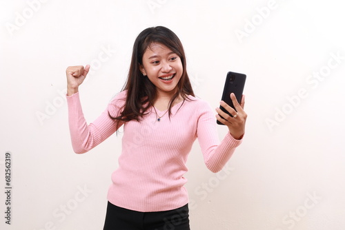 Young asian woman celebrating her victory with fist hand while holding a cell phone. Isolated on white background