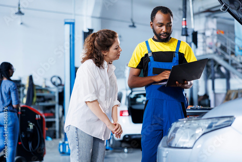 Repairman at auto repair shop conducts yearly vehicle checkup, informing customer about needed engine mending. BIPOC garage worker discussing with customer after finishing car maintenance