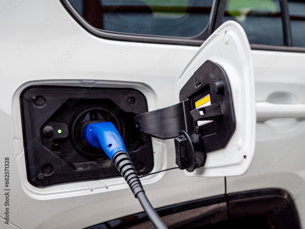 White electric car being charged by cable and plug head. Modern eco friendly car trend. Replacing petrol car with renewable battery agenda. Every day procedure to keep vehicle going White electric car being charged by cable and plug head. Modern eco friendly car trend. Replacing petrol car with renewable battery agenda. Every day procedure to keep vehicle going