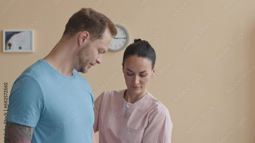 Man raising dumbbell with help of female physiotherapist in clinic while doing rehabilitation exercises after arm injury Man raising dumbbell with help of female physiotherapist in clinic while doing rehabilitation exercises after arm injury