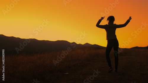 Girl dancing against bright orange sunset sky. Slim young girl silhouette having fun, moving, raise hands up, listening music in mountain field. Back view.