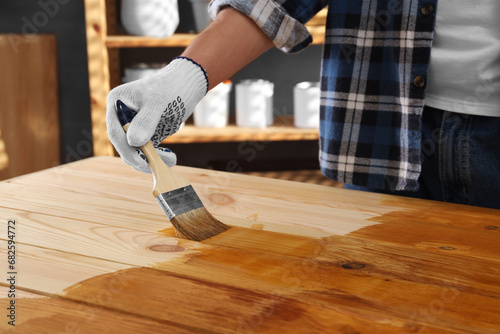 Man with brush applying wood stain onto wooden surface indoors, closeup photo