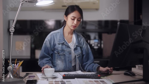 Asian female architect, deeply focused, works on designing products using her computer. She enjoys a cup of hot coffee in a modern office setting, adding to the contemporary ambiance of her workplace photo