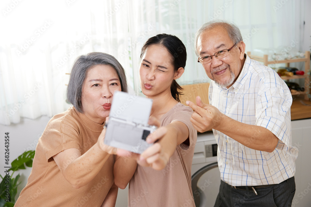 asian family senior couple with granddaughter talking selfie from digital camera, making miniheart sign and duck face at home asian family senior couple with granddaughter talking selfie from digital camera, making miniheart sign and duck face at home