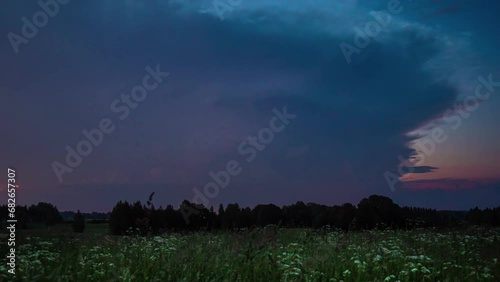 Lightning storm clouds flash light on dark grey day above flower meadow by forest photo