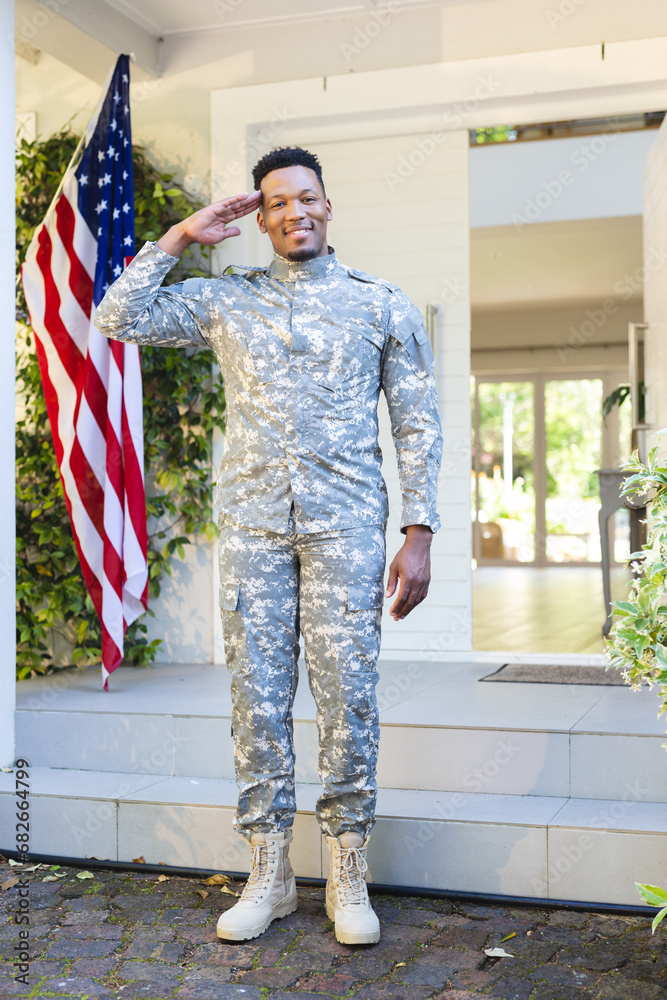 Happy african american male soldier standing and saluting outside home with usa flag, copy space