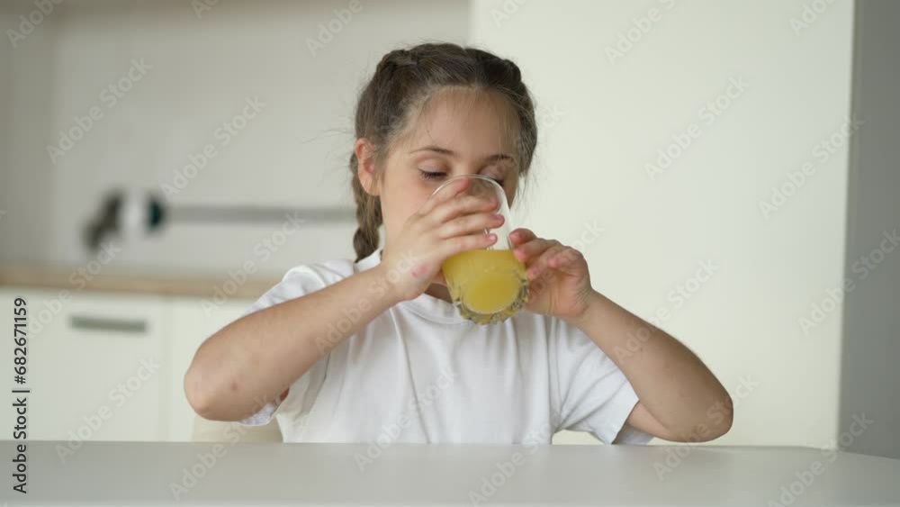 Girl sits at an empty table with a glass of orange juice. Healthy vitamin baby food. Kid with fruit drink in the kitchen. Funny kid drinks juice. Girl healthy food. Child eating breakfast in kitchen