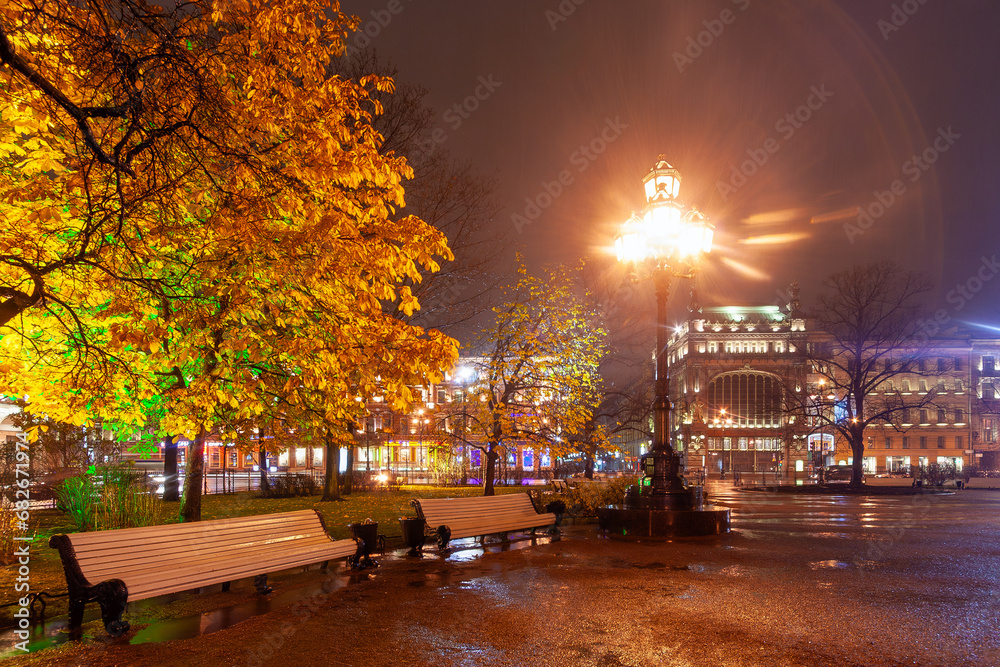 Obraz premium Evening view of empty benches in the city park and beautiful buildings in the historical center of St. Petersburg. Rainy autumn weather. Nevsky Prospekt, St. Petersburg, Russia. Night city.