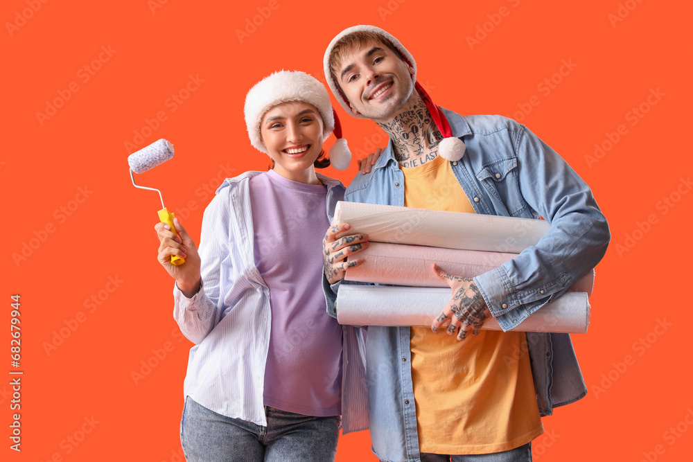 Young pregnant couple in Santa hats with paint roller and wallpapers on orange background