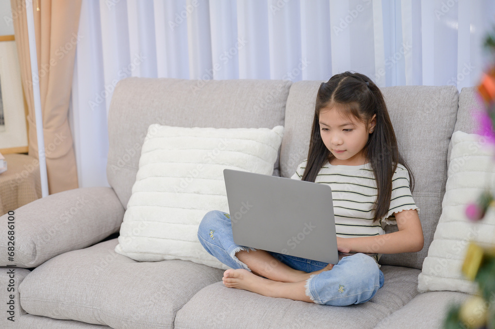 Portrait of a young Asian girl enjoying holiday activities. For education and learning that is fun and happy with using a laptop computer on the sofa in the living room at home Portrait of a young Asian girl enjoying holiday activities. For education and learning that is fun and happy with using a laptop computer on the sofa in the living room at home