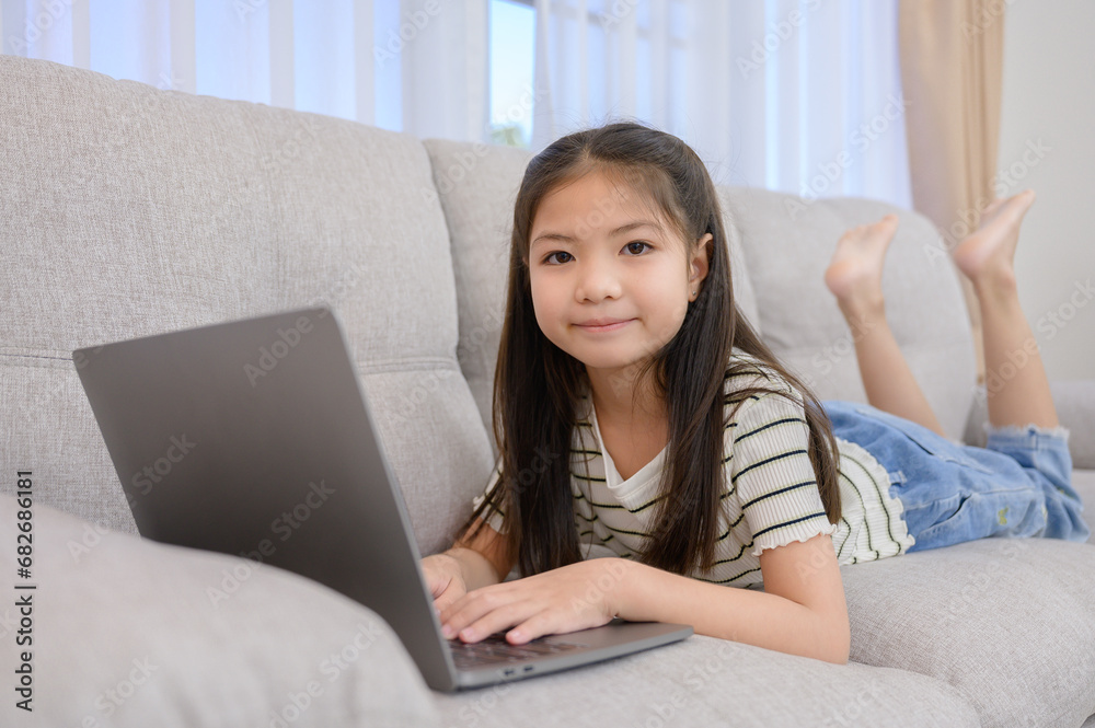 Portrait of a young Asian girl enjoying holiday activities. For education and learning that is fun and happy with using a laptop computer on the sofa in the living room at home Portrait of a young Asian girl enjoying holiday activities. For education and learning that is fun and happy with using a laptop computer on the sofa in the living room at home