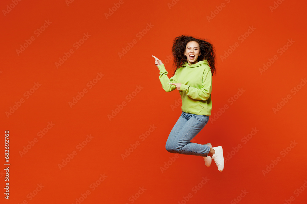 Full body young woman of African American ethnicity she wear green hoody casual clothes jump high point index finger aside on area mock up isolated on plain red orange background. Lifestyle concept. Full body young woman of African American ethnicity she wear green hoody casual clothes jump high point index finger aside on area mock up isolated on plain red orange background. Lifestyle concept.