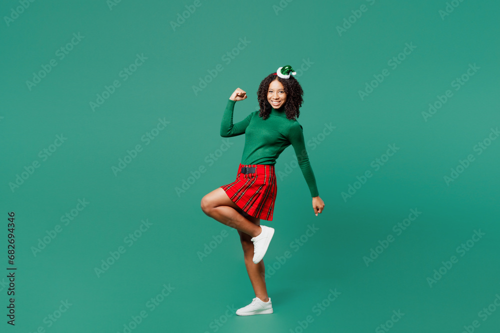 Full body side view merry little kid teen girl wear hat casual clothes posing do winner gesture raise up leg isolated on plain green background. Happy New Year celebration Christmas holiday concept.