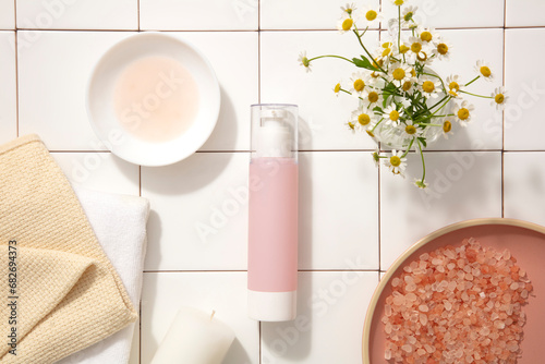 A pastel pink cosmetic bottle with a spray tip is displayed on a white background with pink salt  daisies  towels and plates. Space for product advertising. Top view.