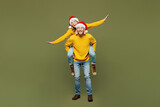 Full body merry young couple two friends man woman wear sweater Santa hat posing giving piggyback ride joyful, sit on back show ok isolated on plain green background. Happy New Year Christmas concept.