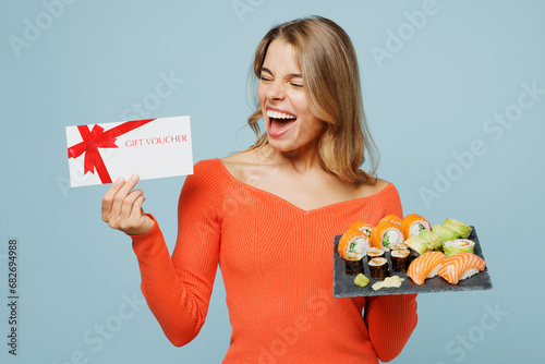 Young happy excited woman wear orange casual clothes hold store gift coupon voucher card eat raw fresh sushi roll served on black plate Japanese food isolated on plain blue background studio portrait.