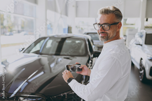 Adult happy man customer male buyer client wear shirt take photo on mobile cell phone choose auto want to buy new automobile in car showroom vehicle salon dealership store motor show. Sales concept.