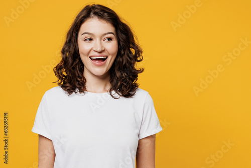 Young surprised happy cheerful fun cool smiling Caucasian woman she wear white blank t-shirt casual clothes looking aside on area mock up isolated on plain yellow orange background. Lifestyle concept.