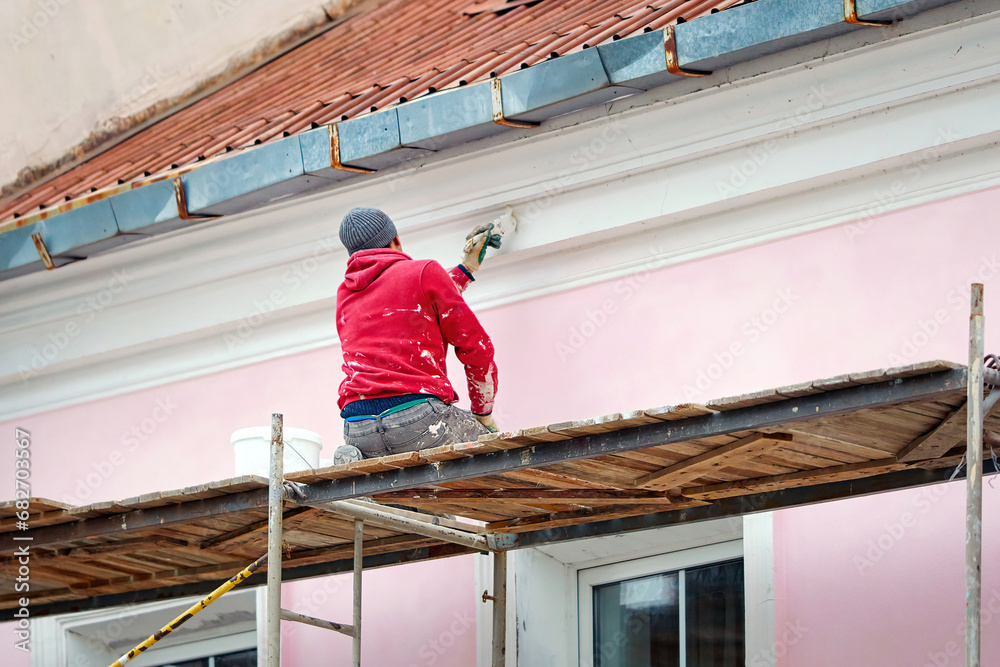 Worker on scaffolding paint facade wall of historic building. Utility worker paints building facade. Repair and restoration building facade. Man painting house wall with paintbrush