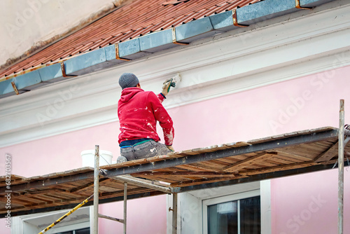 Worker on scaffolding paint facade wall of historic building. Utility worker paints building facade. Repair and restoration building facade. Man painting house wall with paintbrush photo