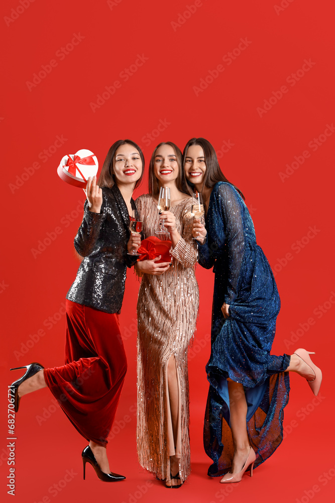Beautiful young women with champagne and gifts on red background. New Year celebration