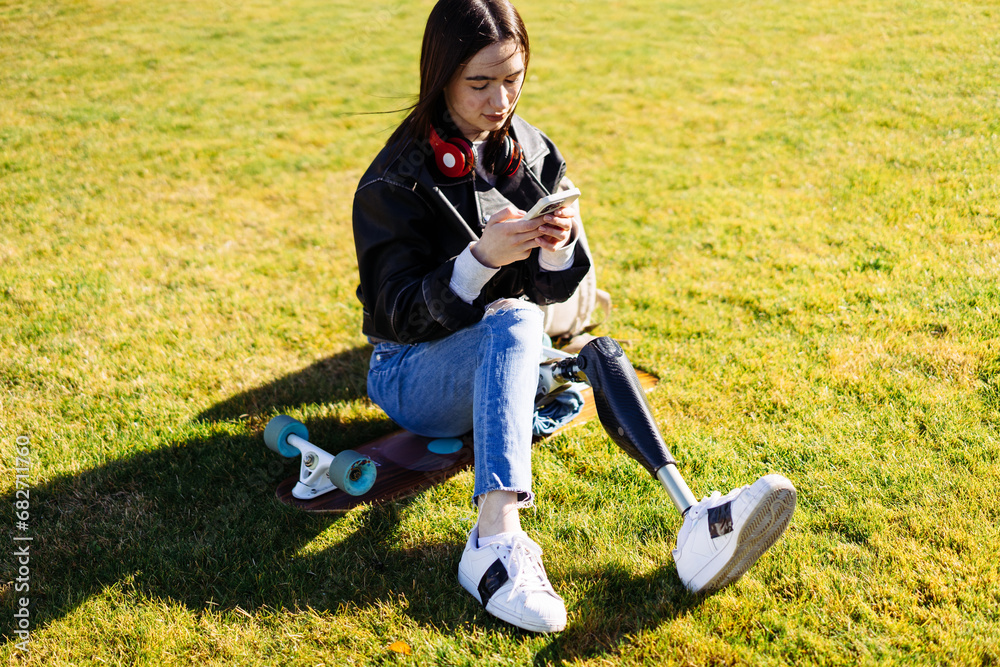 Fototapeta premium Young woman student with prosthetic leg sitting on green grass in university campus and using smartphone. Disabled woman with bionic leg in public park. Woman with prosthesis equipment using phone