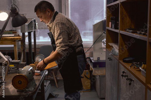 side view portrait of man in checked shirt and black apron standing in front of the table, desk and smoothing the surface of leather cloth