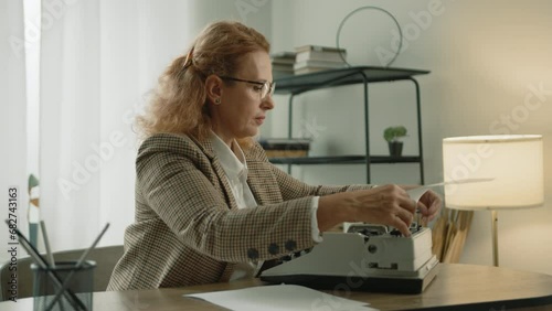 Experienced blonde typist putting sheet of paper into vintage mechanical typewriter, starting typing, sitting at table in light room with modern interior. High quality 4k footage