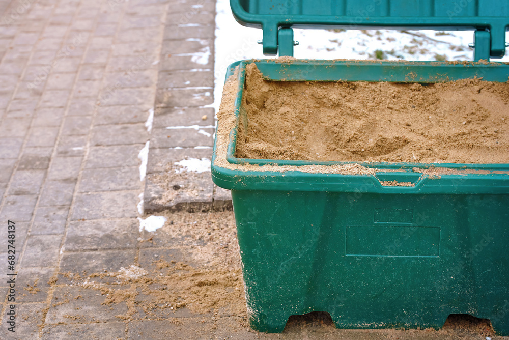 Naklejka premium Grit bin with sand for treatment paving slabs from snow and ice, close up. Sand box for improve traction on snowy, icy sidewalk, road maintenance in winter season. Selective focus