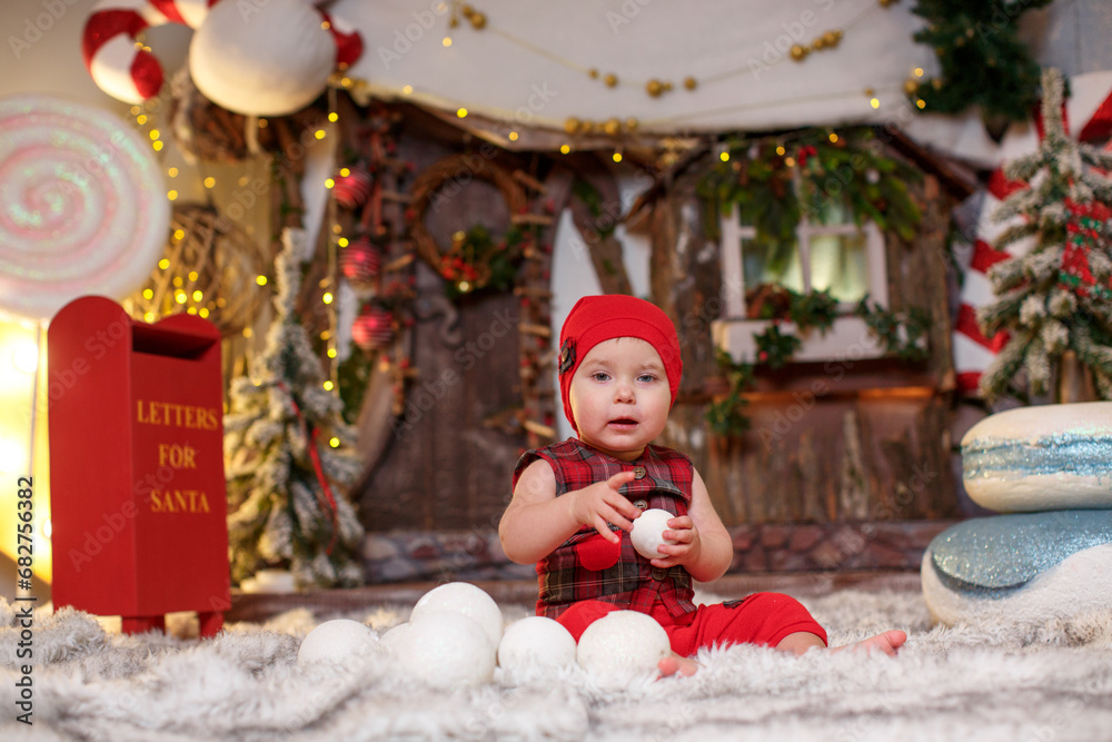 A baby in a checkered vest, a cap and shorts with bare feet in New Year's decorations against the backdrop of a fairy-tale house with Santa's mailbox, huge snowballs and macaroons with caramel