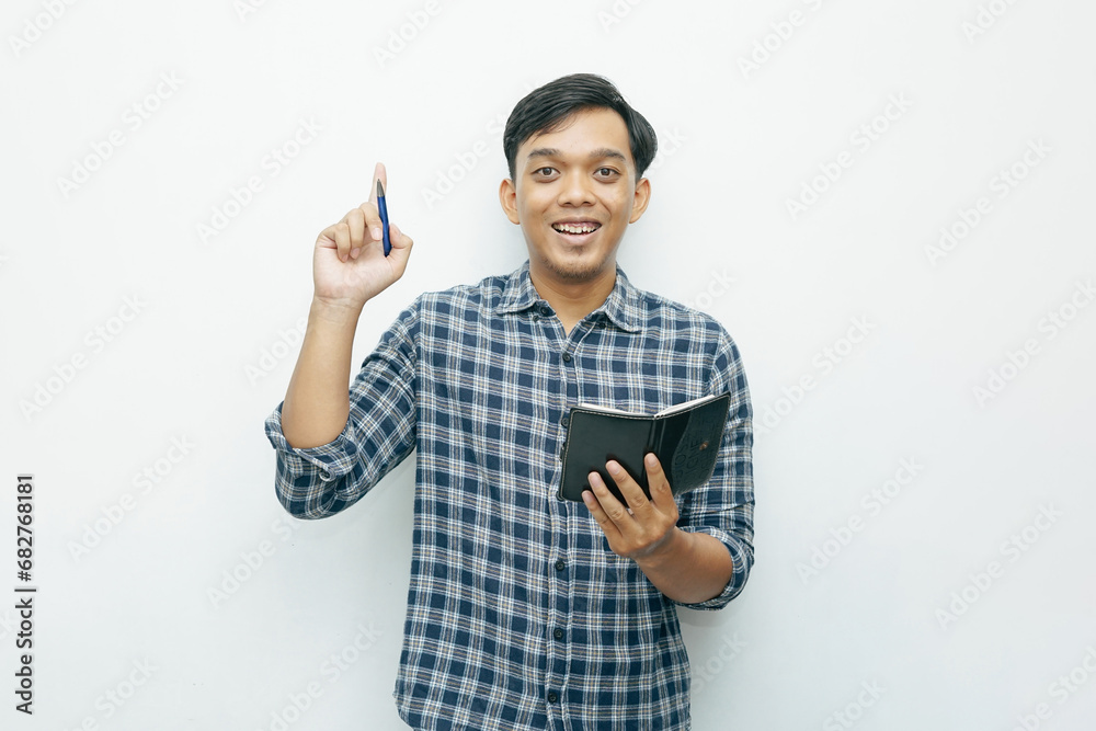 Portrait of young Asian Indonesian man smiling when holding small notebook with ballpoint pen with isolated white background Portrait of young Asian Indonesian man smiling when holding small notebook with ballpoint pen with isolated white background