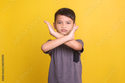 Serious little boy crossing hands and looking at camera with aggression, showing stop gesture over yellow background
