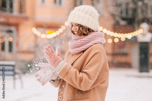 A young beautiful woman shakes snow off her knitted mittens against the backdrop of a winter snow-covered city.