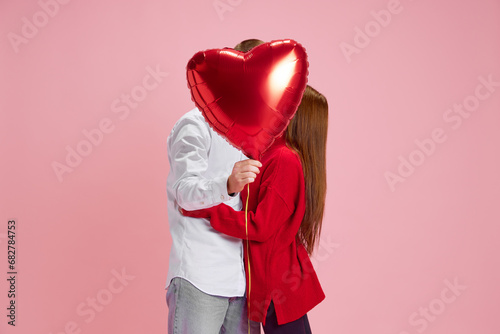 Happy young couple, woman and man holding balloon in heart shape and kissing behind against pink studio background. Concept of love, relationship, Valentine's Day, emotions, lifestyle