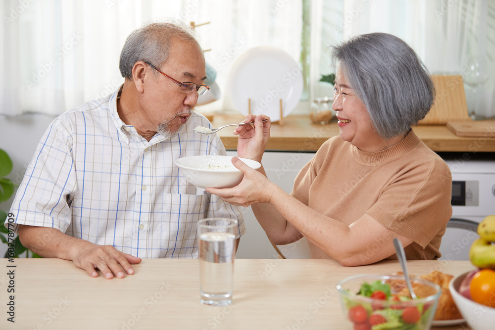 senior woman feeding rice porridge to senior man in the kitchen senior woman feeding rice porridge to senior man in the kitchen