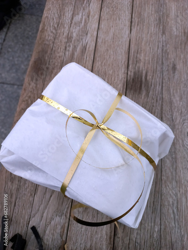 Gift box wrapped in white paper with golden ribbon on a wooden background under the rain