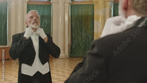 Medium over shoulder shot of mature Caucasian man with grey hair and beard standing in front of mirror in classical banquet hall and adjusting vest and tie with gloved hands photo