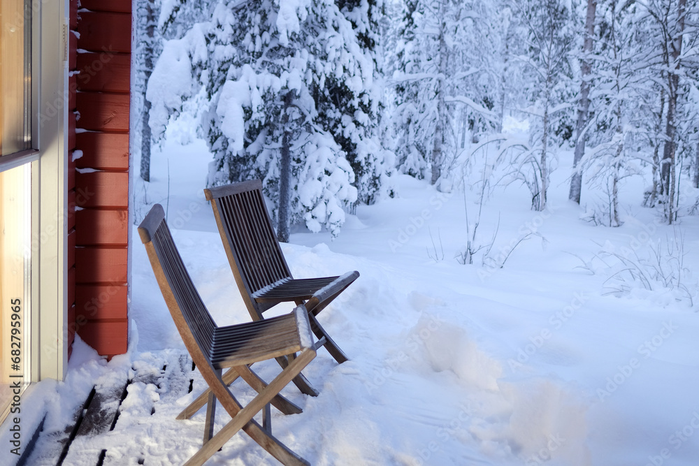 early evening in winter forest, cozy wooden chairs on terrace of country house, solid light pours from large windows, snow-covered trees, concept of winter holiday, weekend, new year in Lapland