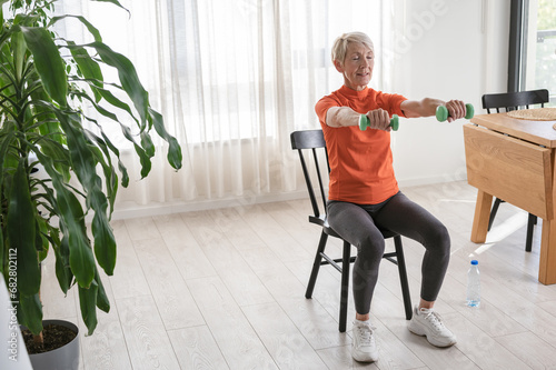 Smiling beautiful senior woman health instructor doing chair exercises with dumbbells