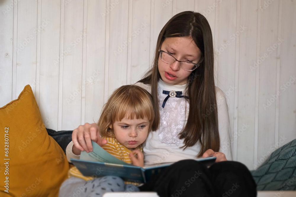 Teenager Girl reading book to her curious little sister baby girl at home. Close up. Native sisters spending time together. Family time comes in various forms. Two girls read story in a bedroom. Teenager Girl reading book to her curious little sister baby girl at home. Close up. Native sisters spending time together. Family time comes in various forms. Two girls read story in a bedroom.