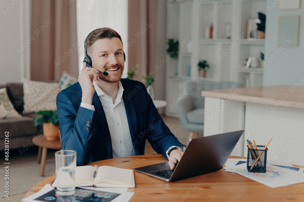 Smiling businessman in headphones sitting at desk at home office and working on laptop Smiling businessman in headphones sitting at desk at home office and working on laptop
