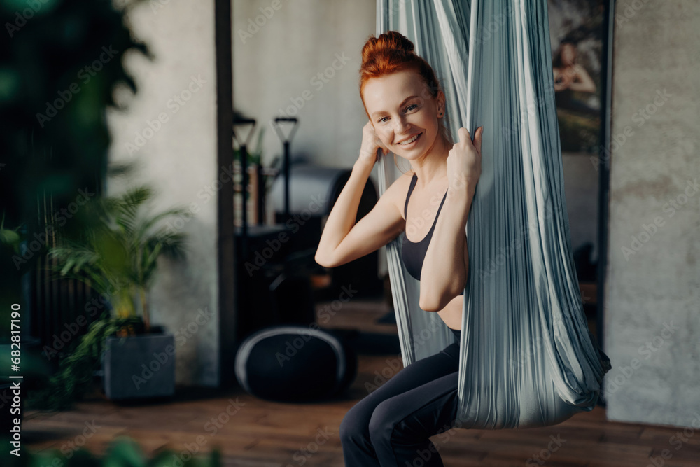 Naklejka premium Happy red haired woman sitting in aerial yoga hammock