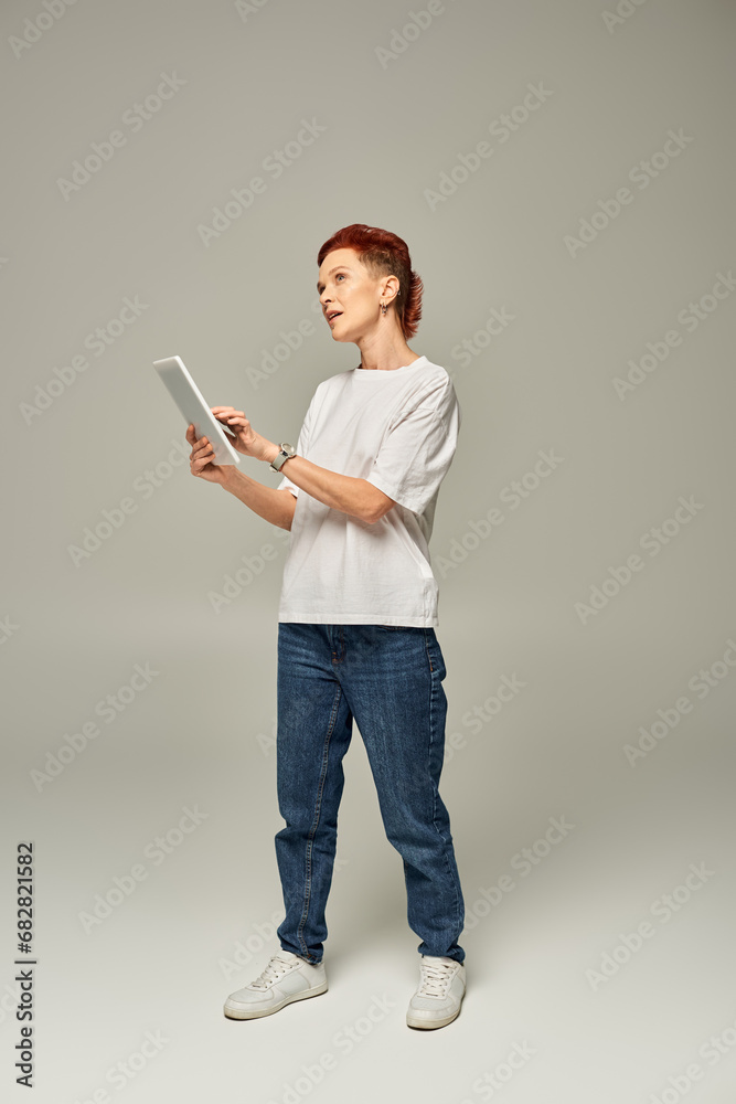 thoughtful non-binary person in white t-shirt using digital tablet and looking away on grey backdrop thoughtful non-binary person in white t-shirt using digital tablet and looking away on grey backdrop