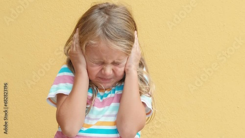 Unhappy sad angry little child girl close eyes and cover ears by hands being annoyed with loud noise, isolated yellow studio background. Children emotions. Child dislike loud sounds photo