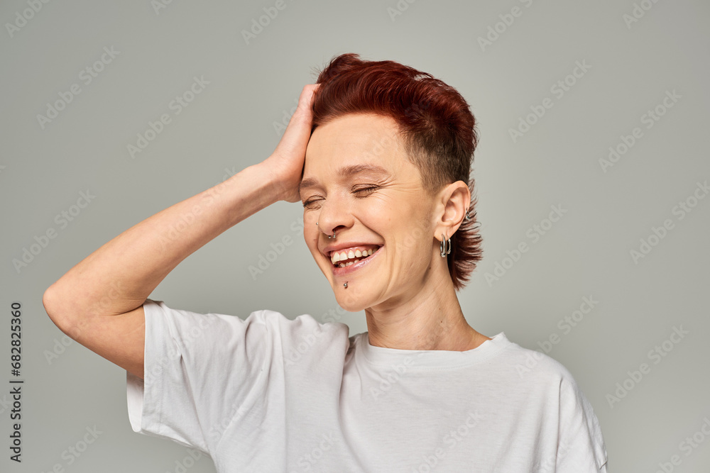 cheerful non-binary person in white t-shirt touching red hair and smiling with closed eyes on grey cheerful non-binary person in white t-shirt touching red hair and smiling with closed eyes on grey